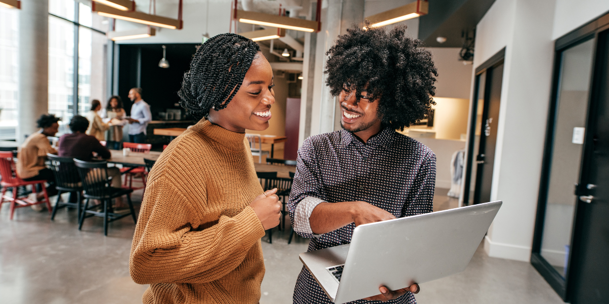 Two people are stood in an office. The man is holding a laptop and pointing at the screen while the woman looks on.