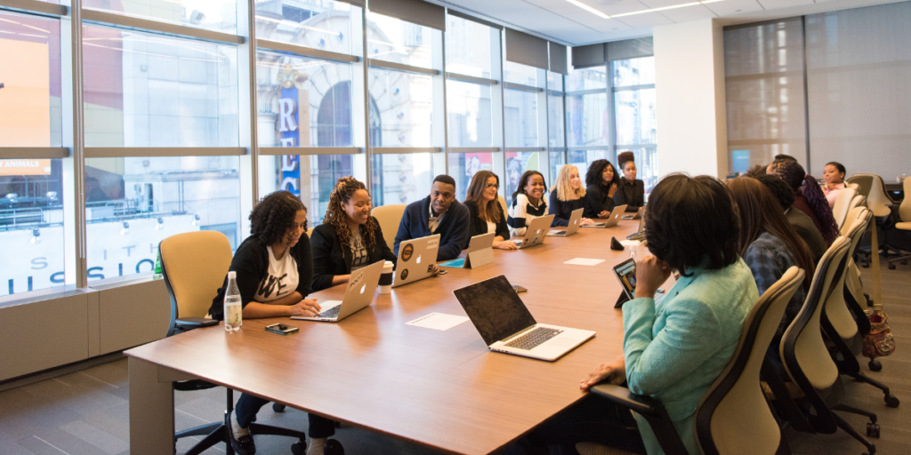 A boardroom full of people sitting at the table while a woman stands at the whiteboard presenting.