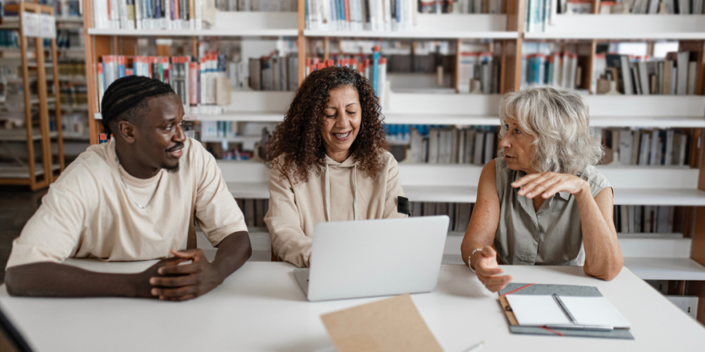 Three people are sat at a desk in a library. One woman is typing as they all chat.