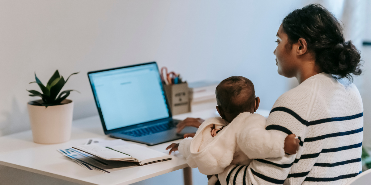 A woman holding a baby is sitting at a desk typing.