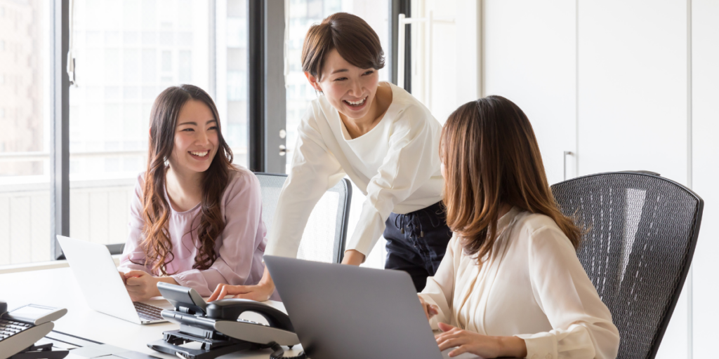 Three women in an office are conversing. Two women are sat with computers and the other is standing.