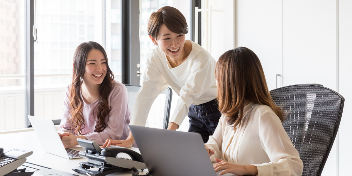 Three women in an office are conversing. Two women are sat with computers and the other is standing.