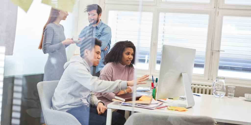 Four people are in an office and two are sat at a desk looking at a monitor.