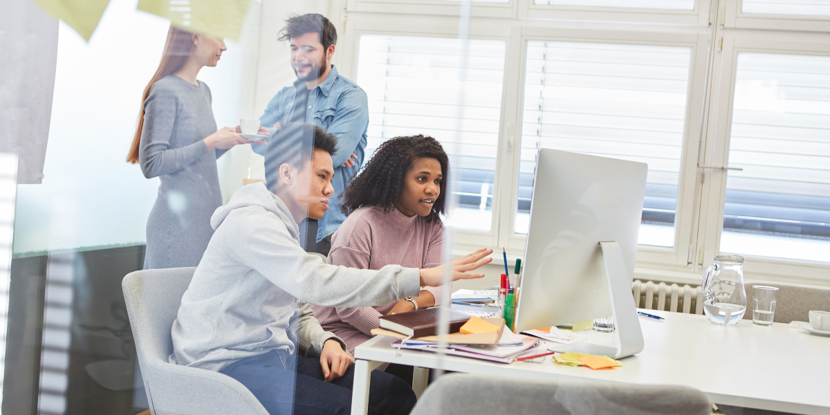 Four people are in an office and two are sat at a desk looking at a monitor.