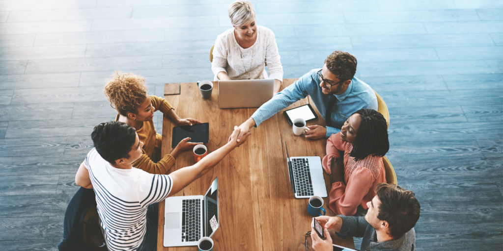 A group of people sat around a boardroom table. Two men are shaking hands across the table.