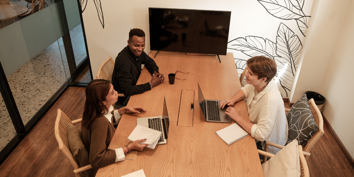 Three people sitting at a boardroom table in a dimly lit room. Two of the individuals have their laptops and they are all conversing.