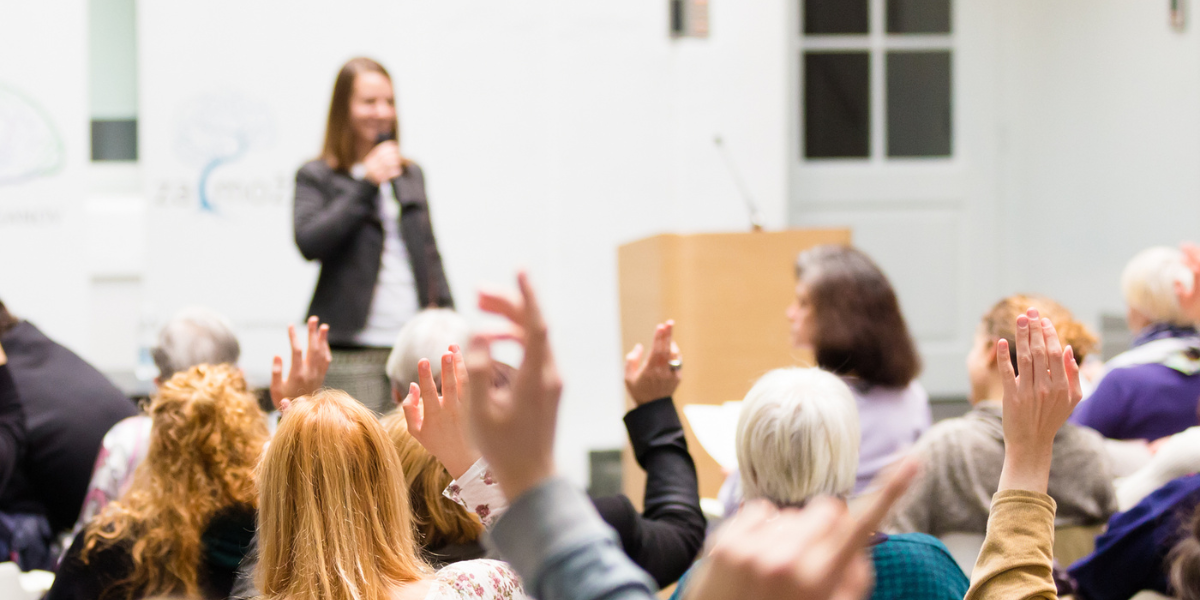 A room full of conference goers with hands raised and the presenter is standing at the front of the room.