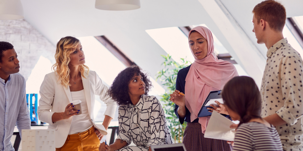 A large group of individuals are surrounding a boardroom table in a white dimly lit room. One of the women is stood holding a tablet and presenting to the team.