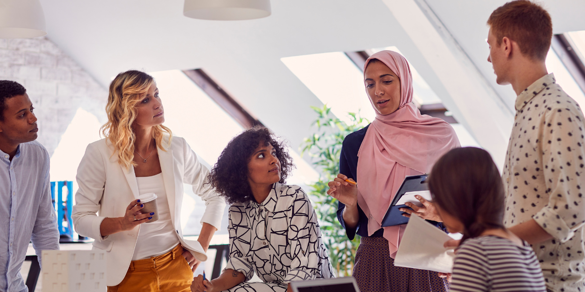 A large group of individuals are surrounding a boardroom table in a white dimly lit room. One of the women is stood holding a tablet and presenting to the team.