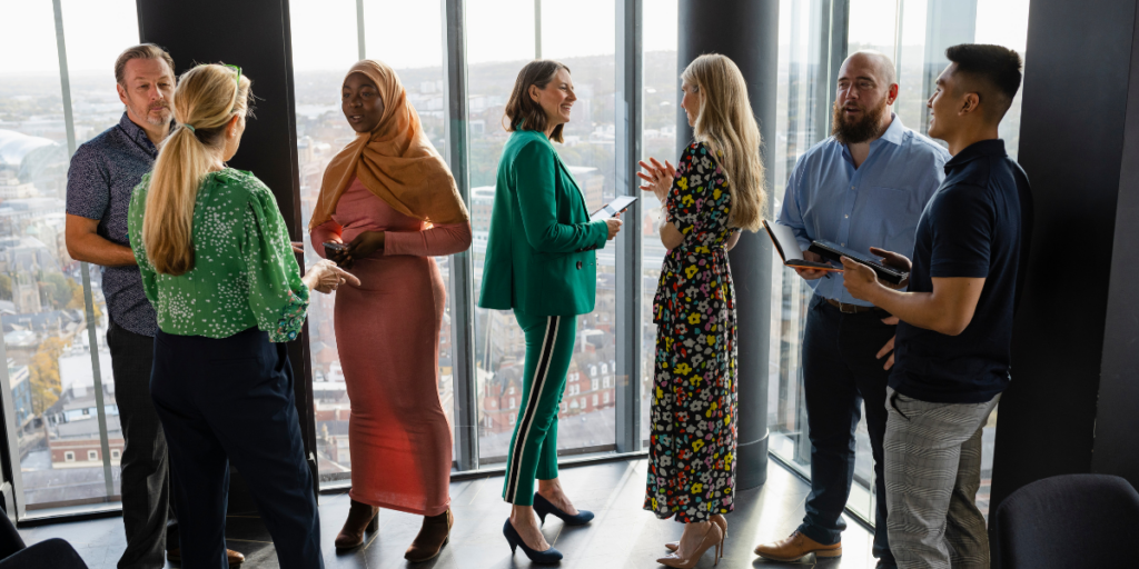 7 people standing in front a wall of windows conversing and 3 of the individuals are holding tablets appearing to interview three of the individuals.