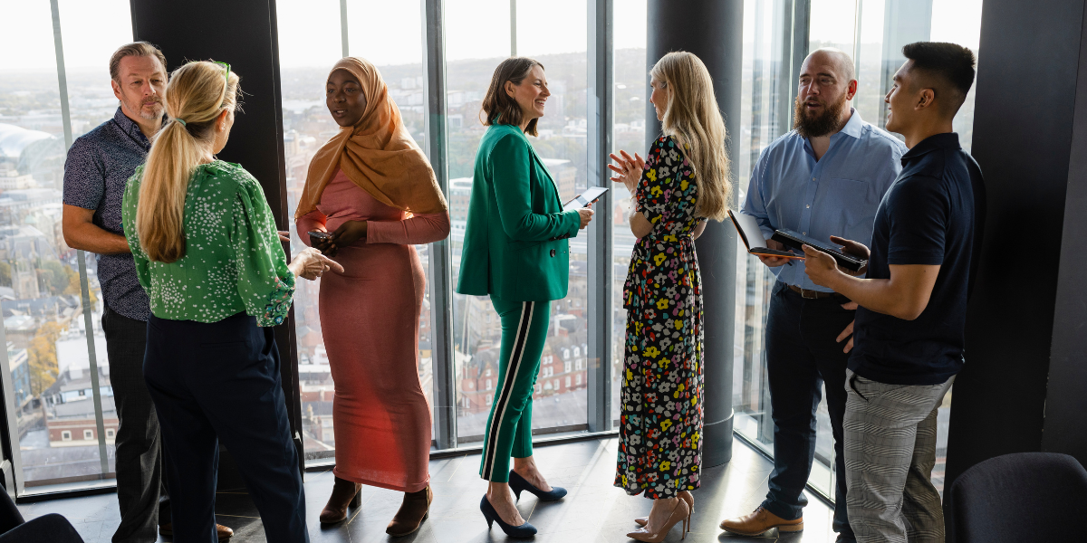 7 people standing in front a wall of windows conversing and 3 of the individuals are holding tablets appearing to interview three of the individuals.