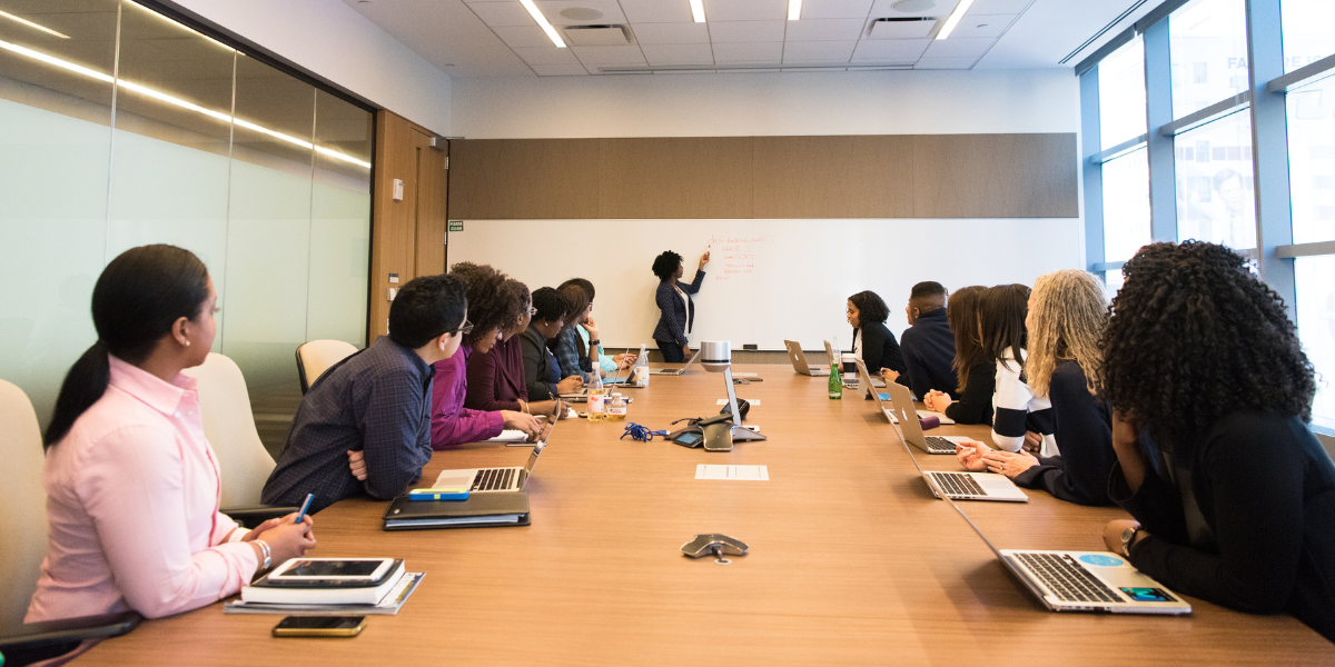 A boardroom full of people sitting at the table while a woman stands at the whiteboard presenting.