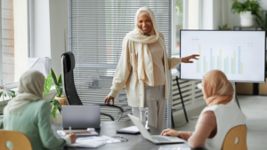 A woman standing at the front of the boardroom presenting statistics from a screen to two of her colleagues.