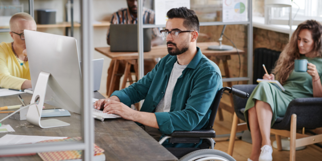 A man in a wheelchair sitting at a dark brown desk typing on a desktop computer. A woman sits in the back writing on a notepad while drinking coffee.