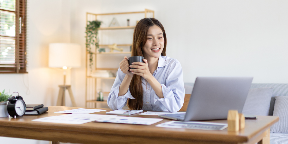 A woman is sat at a wooden desk holding a coffee mug and looking at her laptop.