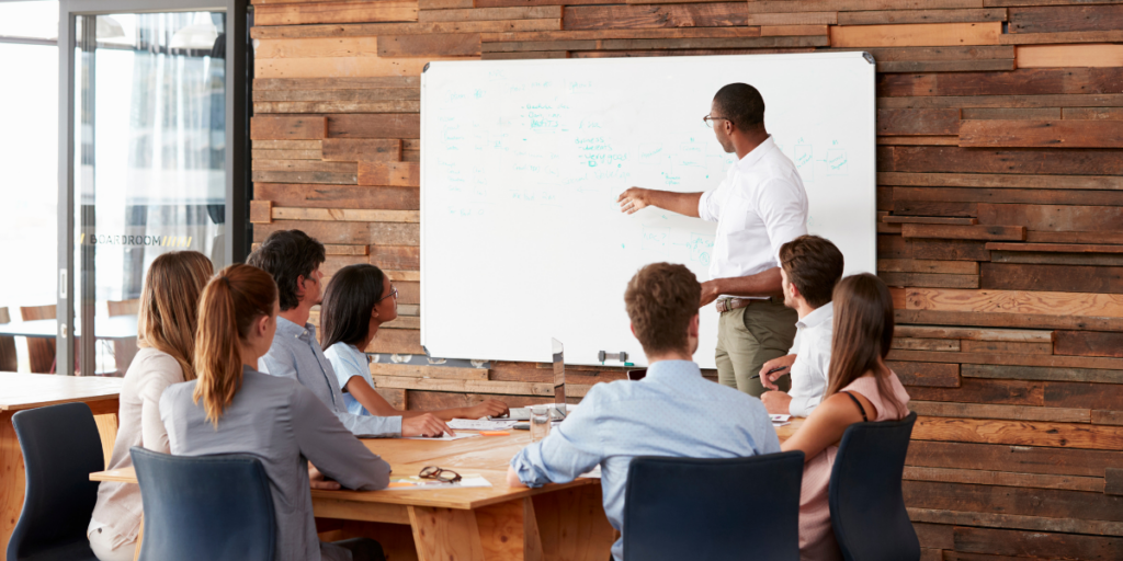 A man is stood at the front of a boardroom next to a whiteboard featuring statistics. The team members sat at the boardroom table are sat facing the whiteboard.