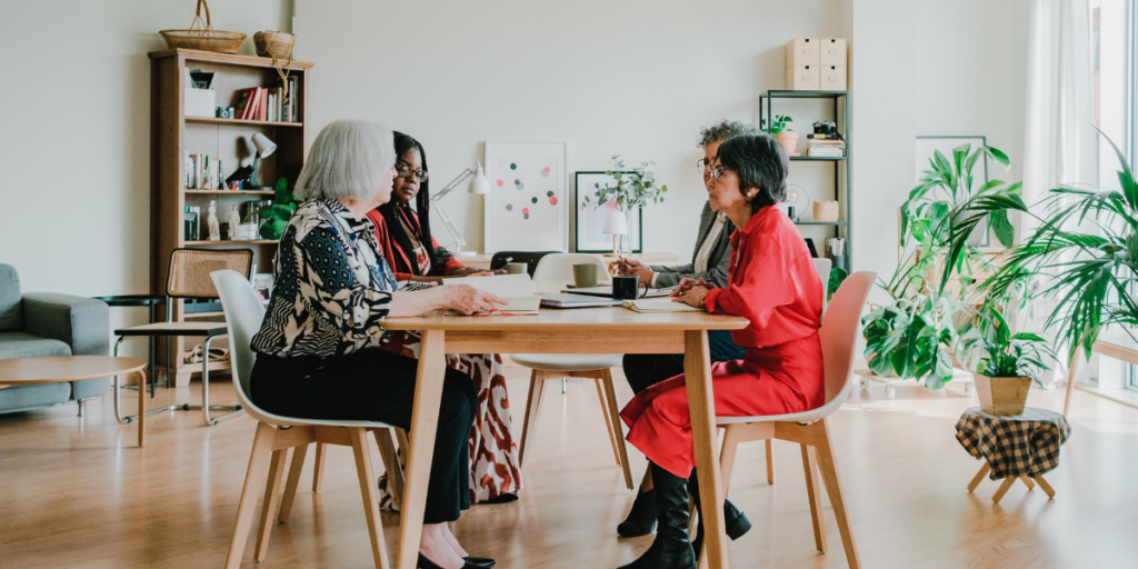A group of women are sat at a boardroom table conversing in a well-lit room with plants.