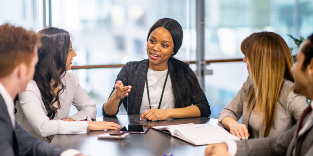 A woman is sat at the head of a boardroom table speaking to a group of employees.
