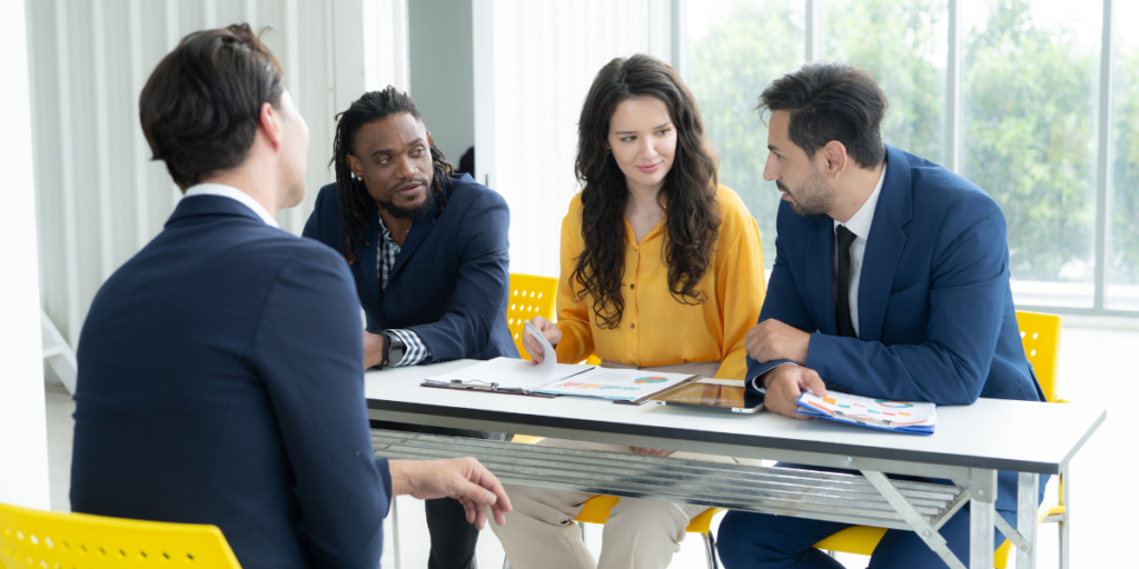 A panel of three employees is interviewing a job candidate in a well-lit office boardroom.