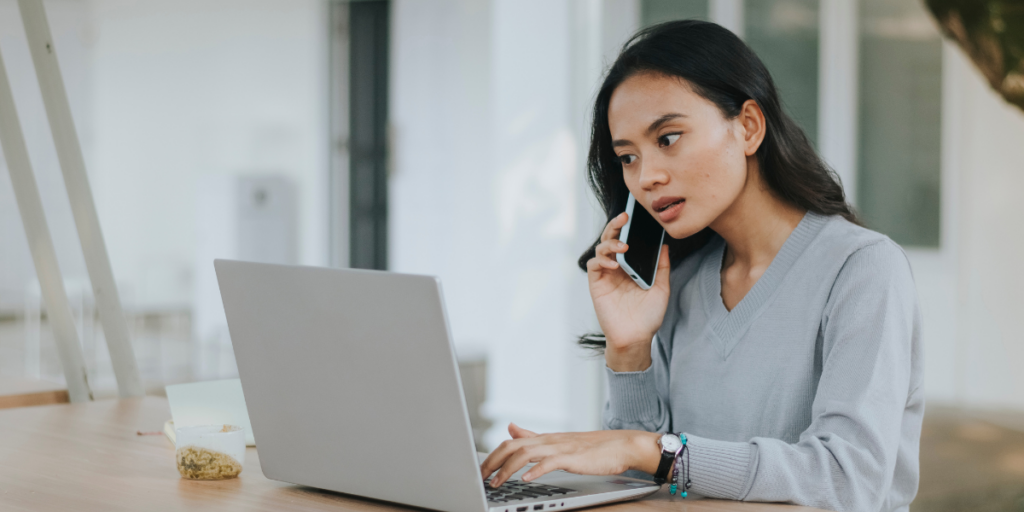 A woman is sat at a desk typing on a laptop and holding a cellphone to her ear.