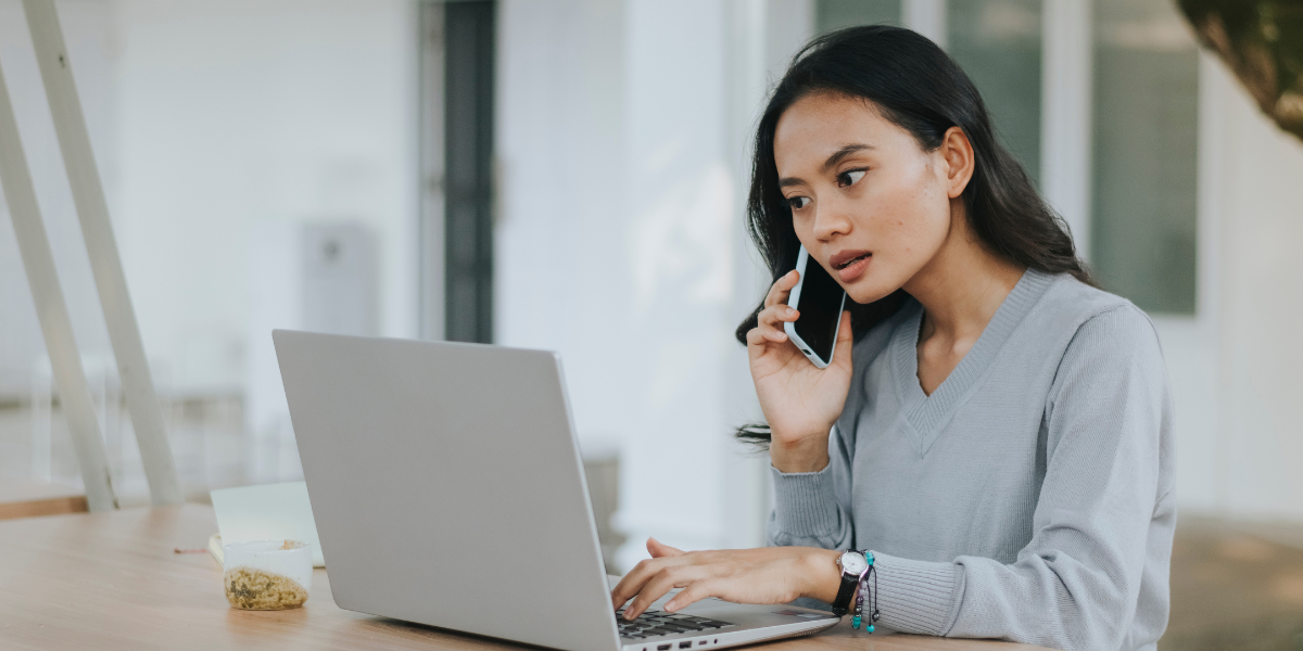 A woman is sat at a desk typing on a laptop and holding a cellphone to her ear.