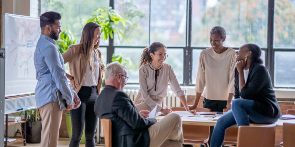 A group of six employees is sat at a boardroom table in front of a wall of windows conversing.