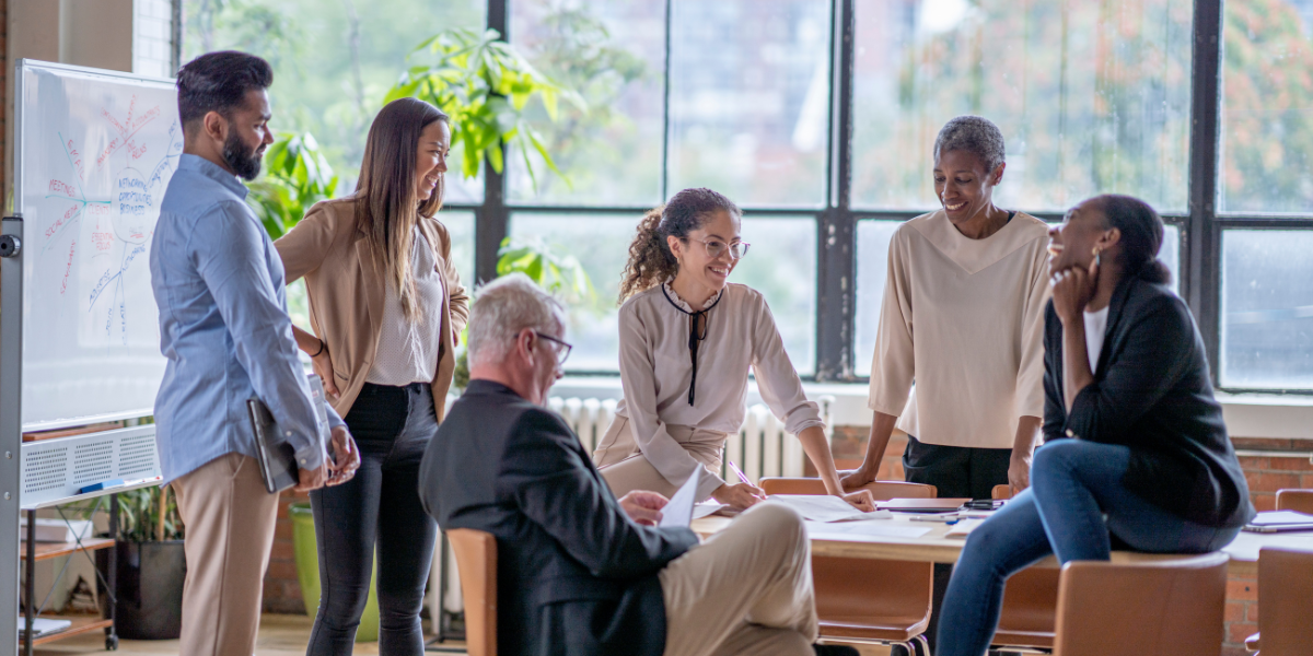 A group of six employees is sat at a boardroom table in front of a wall of windows conversing.