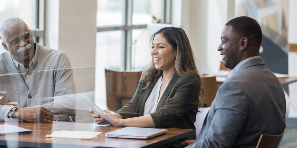 Three people are sat at a boardroom table conversing. The woman is laughing and holding a piece of paper.