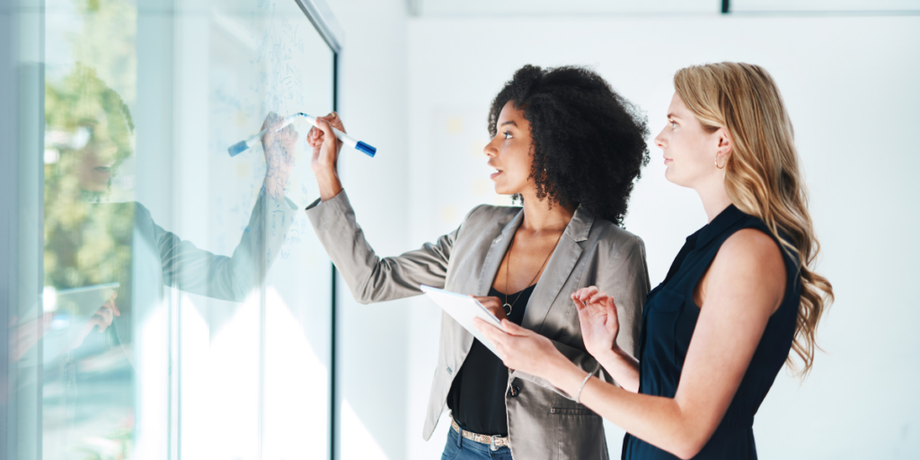 Two women are stood side-by-side writing on a whiteboard, and taking notes.