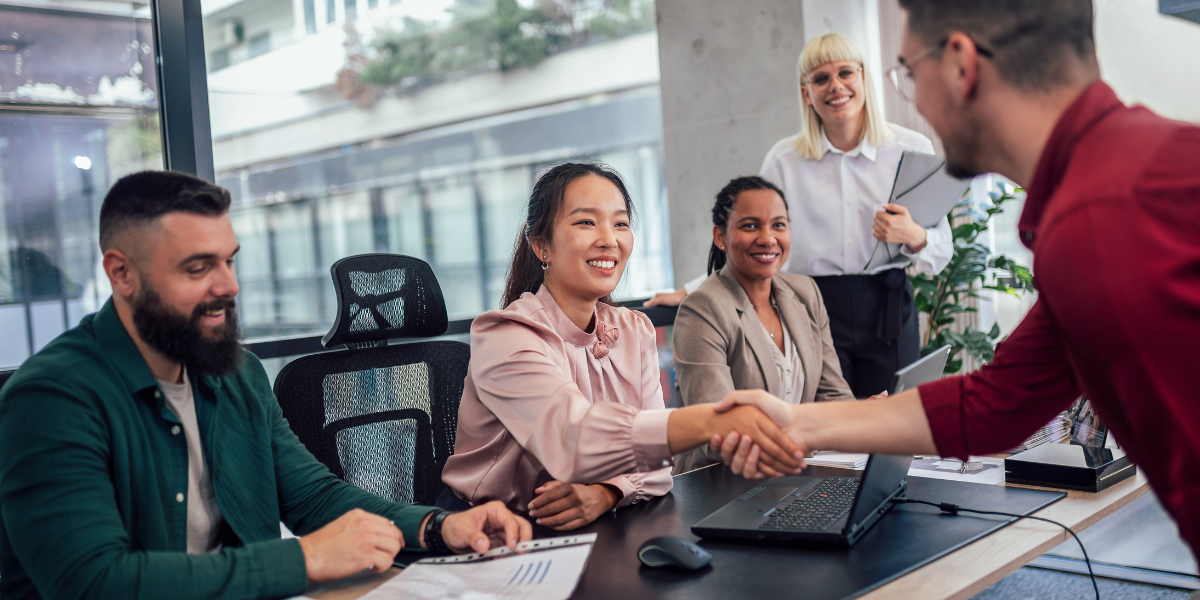Four people are situated around a boardroom table, one woman is shaking hands with a man across the table.