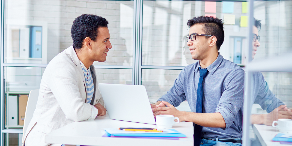 Two men are sat at a desk having a meeting, and there is also a laptop open on the desk.