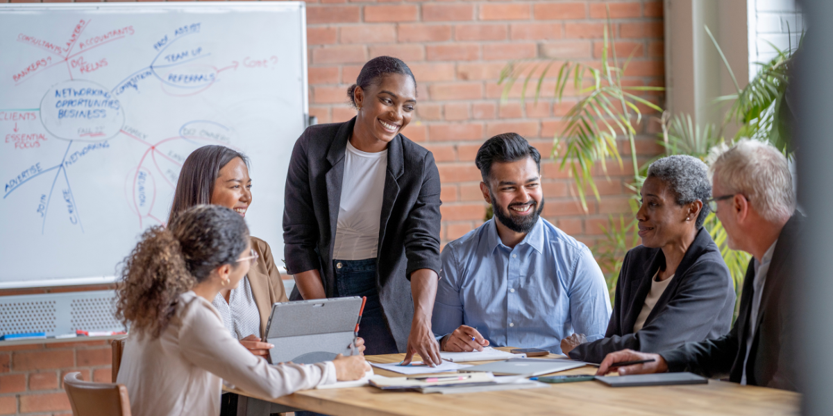 A group of individuals are sat at a boardroom table in a bright room. A woman stands at the front of the table presenting.
