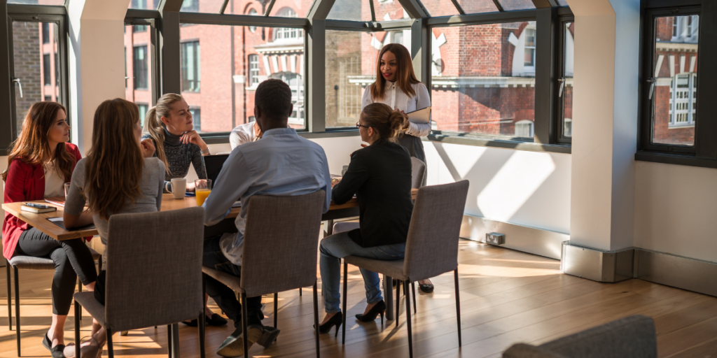 A woman standing at the front of the boardroom presenting to six of her colleagues.