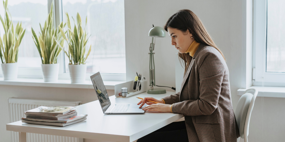 A woman in a brown blazer is sat at a white desk working on a latop.
