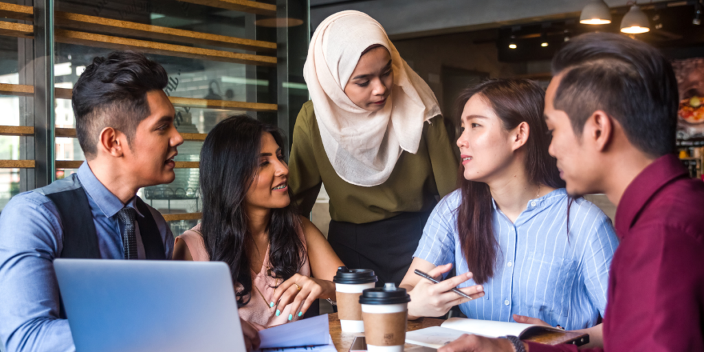 A group of five people are sat and standing around a table conversing and working.