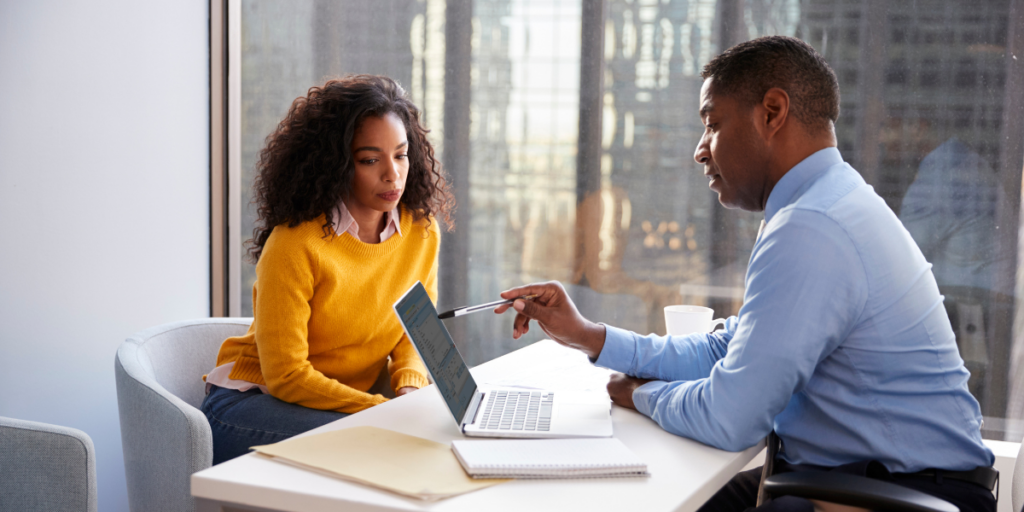 A woman is sat on one side of the desk and a man is sat on the other facing a laptop and discussing metrics.