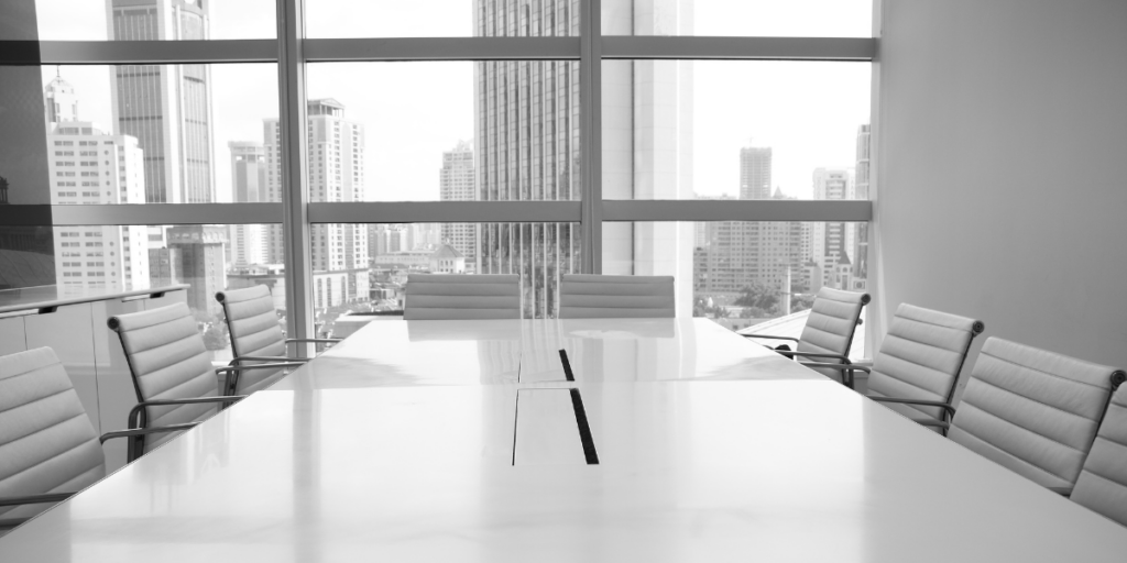 A boardroom with a set of white chairs surrounding a white table with a wall of windows as the backdrop.