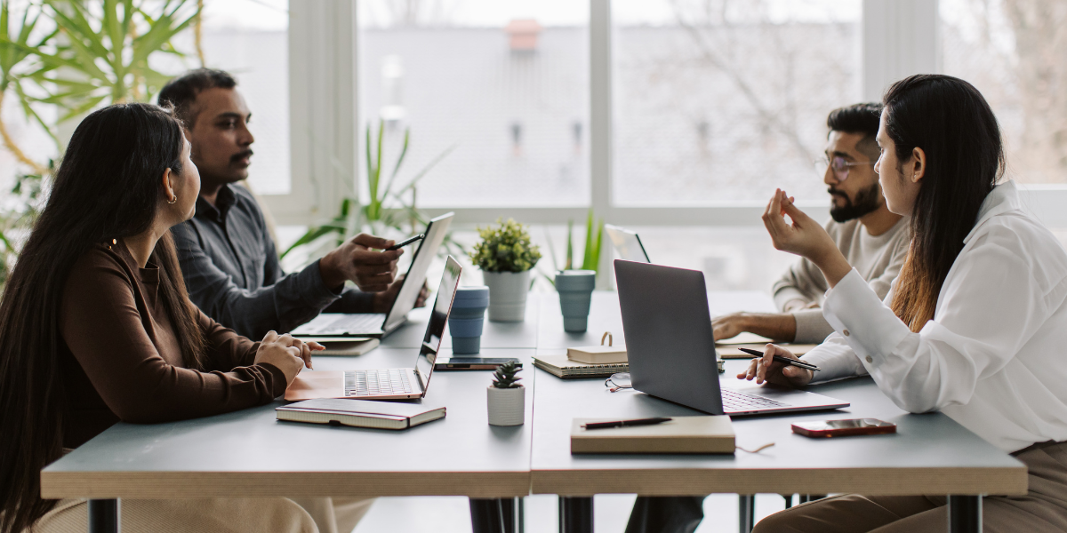 Four people are sat at a boardroom table conversing and working.