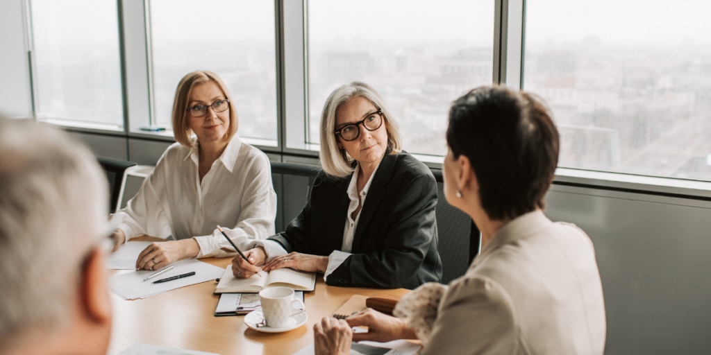 A group of individuals are sat at a boardroom table in a bright room with windows overlooking the city.