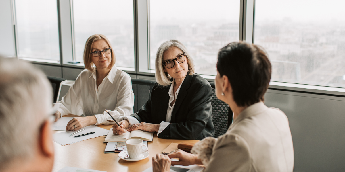 A group of individuals are sat at a boardroom table in a bright room with windows overlooking the city.