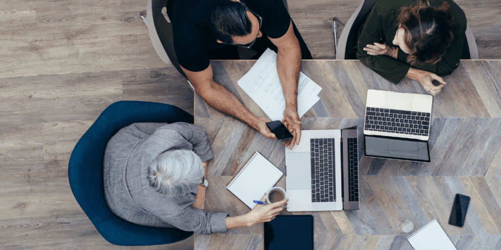 Three people are sat at a boardroom table working and conversing.