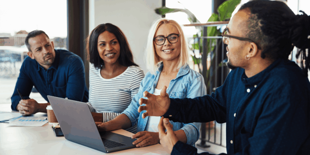 Four people are sat at a boardroom table in a well-lit office space conversing.