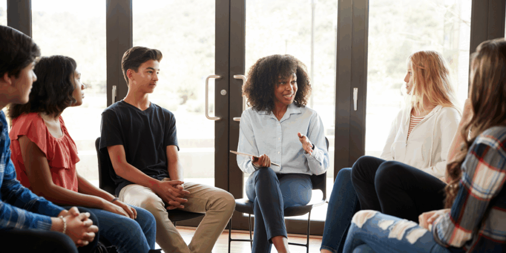 A woman is sitting in front of set of doors presenting to a group of young people all sat in a circle.