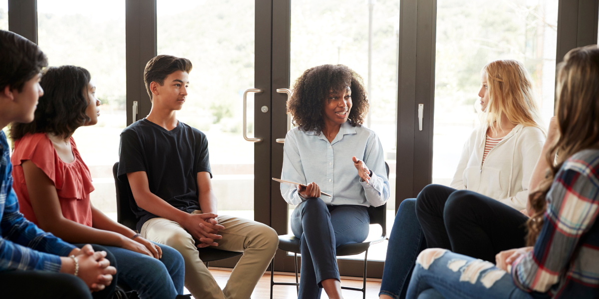 A woman is sitting in front of set of doors presenting to a group of young people all sat in a circle.
