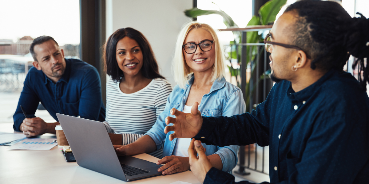 Four people are sat at a boardroom table in a well-lit office space conversing.