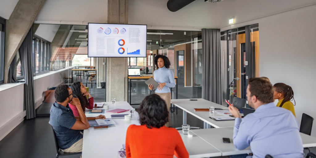 A boardroom of six people sat at boardroom tables in a U-shape with a woman stood at the front presenting.