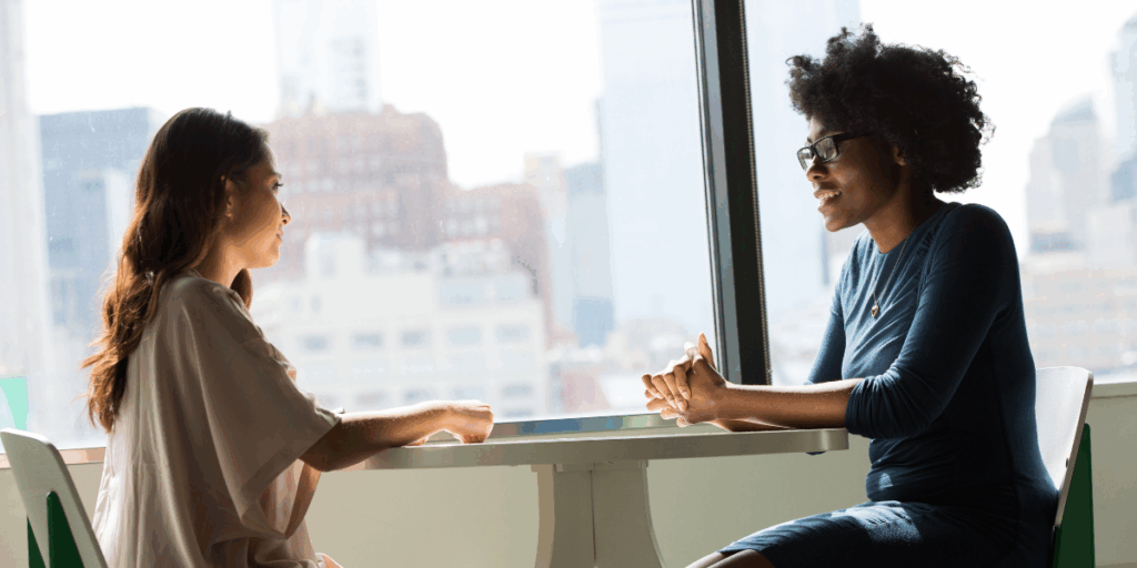 Two women sat at a table in front of a set of windows conversing.