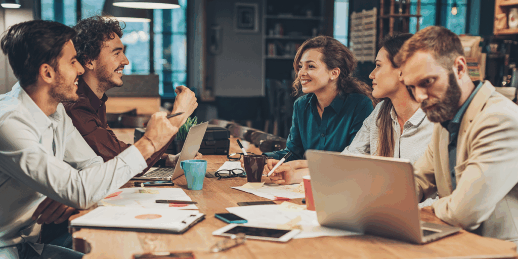 Five people are sitting at a boardroom table with papers strewn on the table. One woman is standing presenting and there is a whiteboard behind with writing on it.