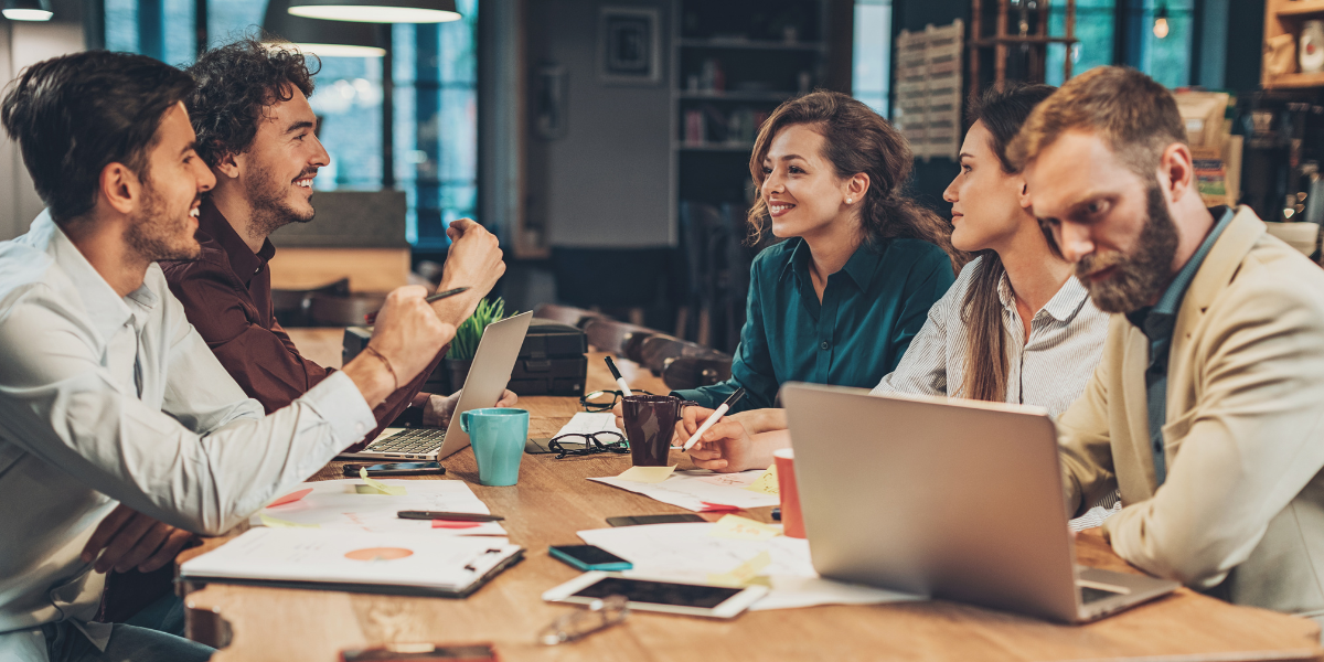 Five people are sitting at a boardroom table with papers strewn on the table. One woman is standing presenting and there is a whiteboard behind with writing on it.