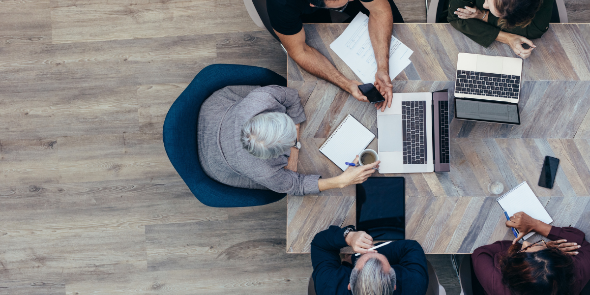 Five people are sitting at a boardroom table working on laptops and conversing.